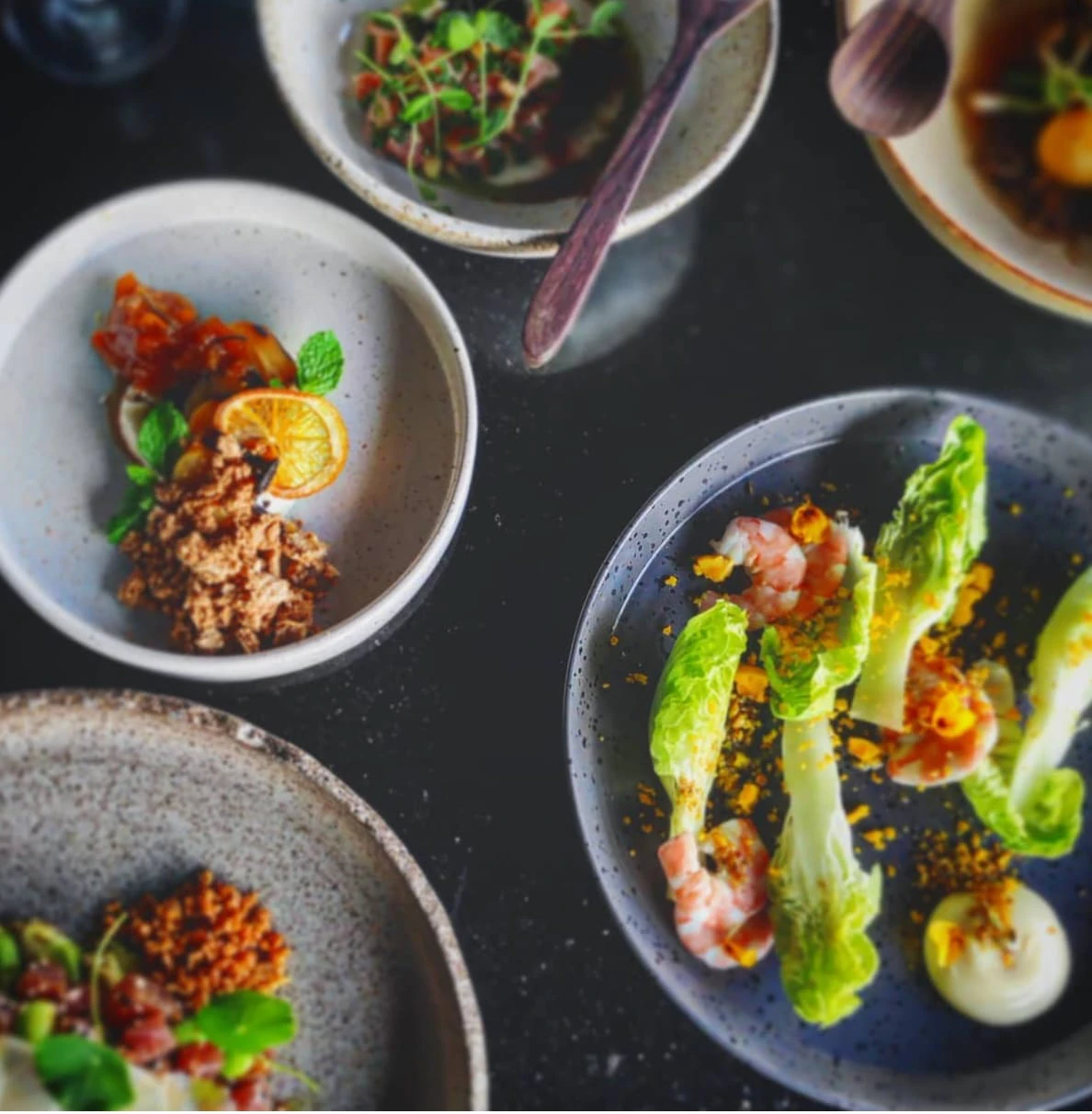 Several small plates of prawns, tartare and garnishes on a dark surface