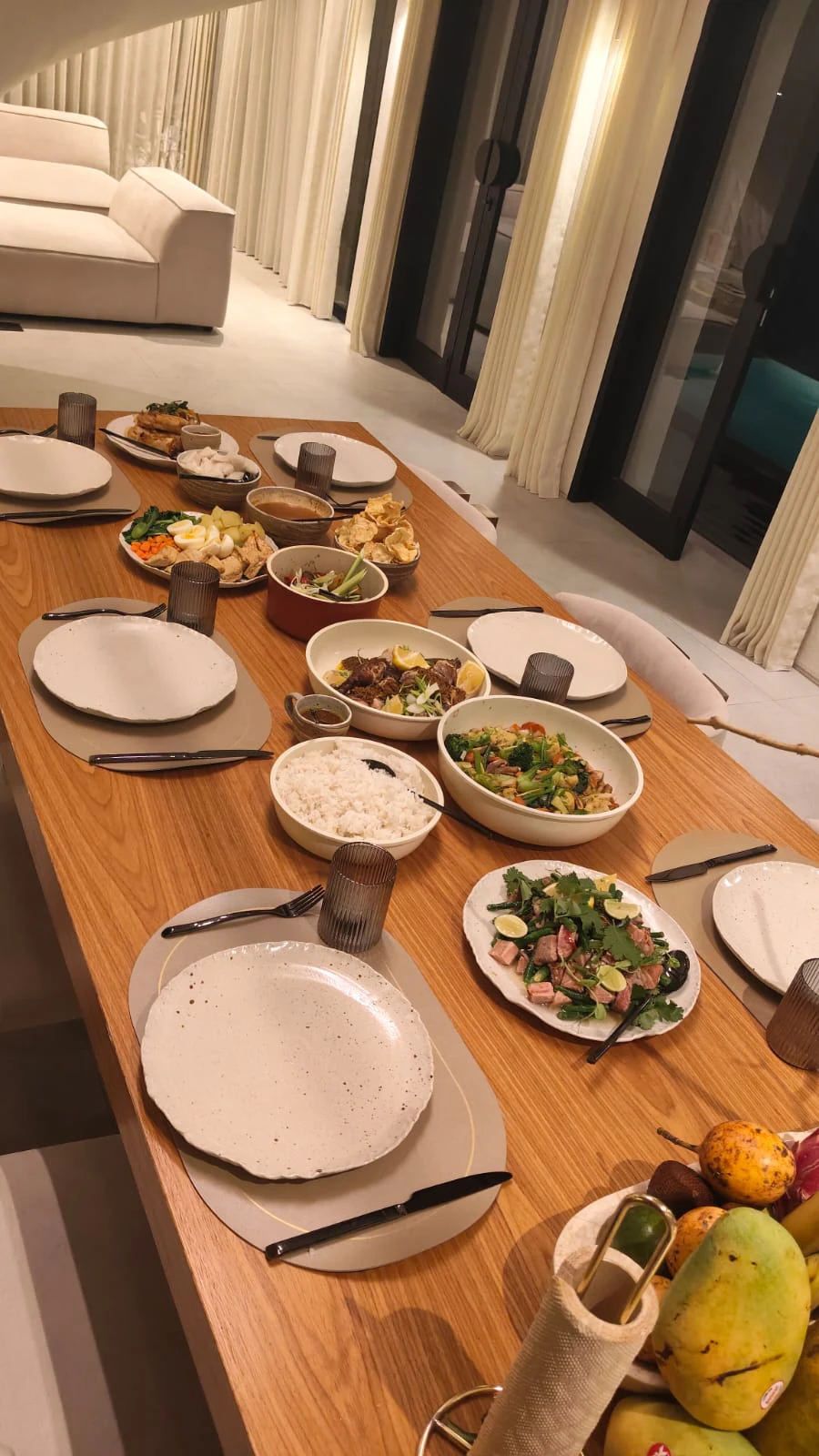 A long dining table laid for dinner inside a Bali villa, warm lamps and linen curtains behind it